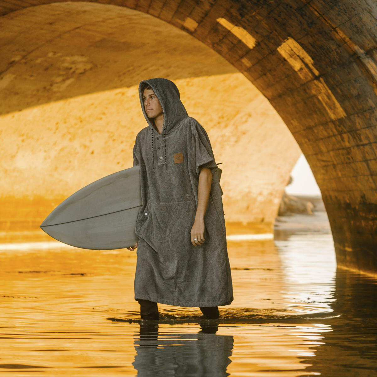 Gray cotton terry towel poncho with surfboard, shown outdoors under a sunlit bridge.
