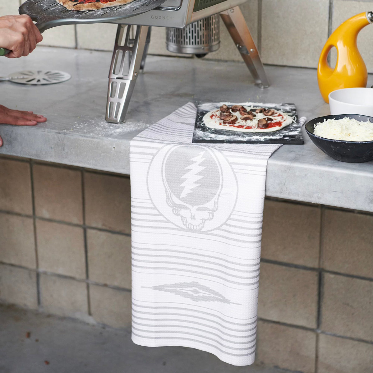Kitchen towel with stealie logo design next to pizza oven, pizzas, and cheese.
