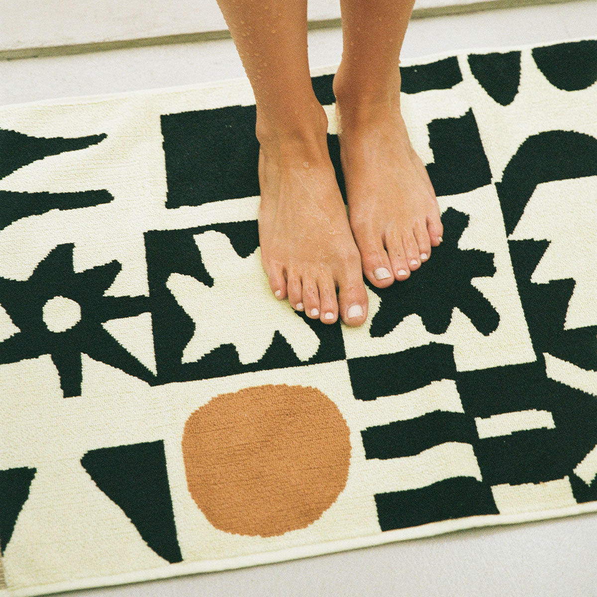 Bath mat with abstract black shapes and brown circle on cream background.