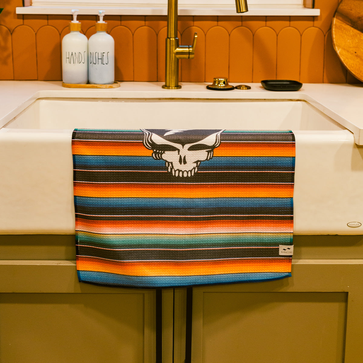 Vintage cotton kitchen towel with colorful stripes and skull logo over a white farmhouse sink.