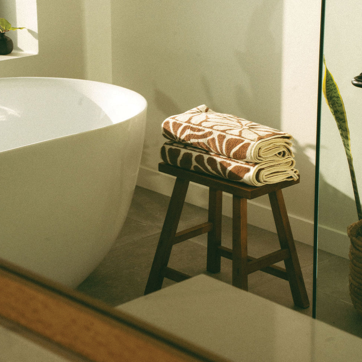 Bath towels with a brown leaf pattern stacked on a wooden stool by a bathtub.