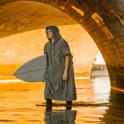 Gray cotton terry towel poncho with surfboard, shown outdoors under a sunlit bridge.