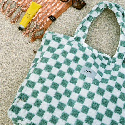Tote bag with checker pattern on sand with towel, sunglasses, and sunscreen nearby.