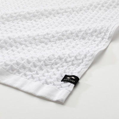 Waffle textured white bath towel with small black corner tag on white surface.
