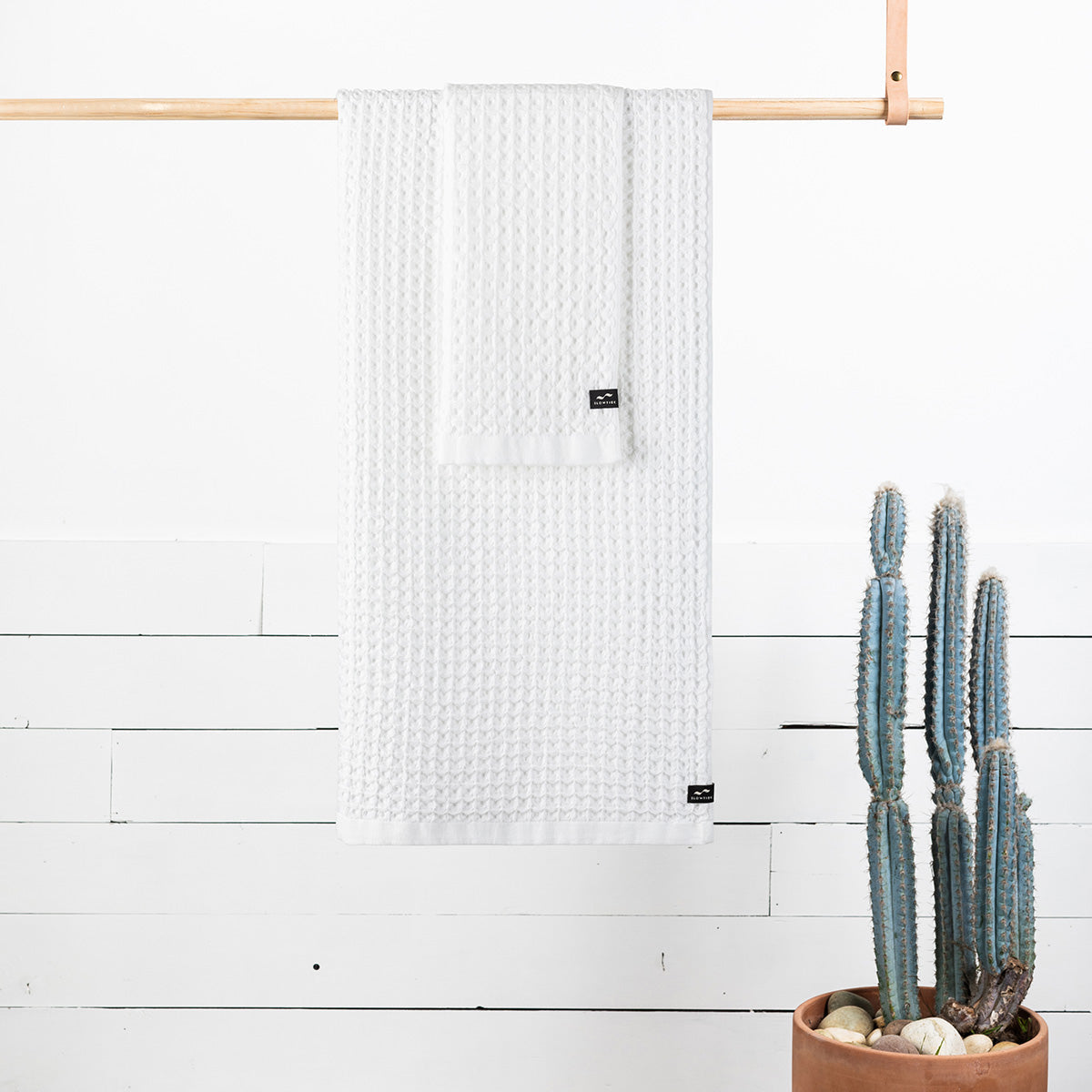 White waffle bath towels on wooden rod above potted cactus near white wall.