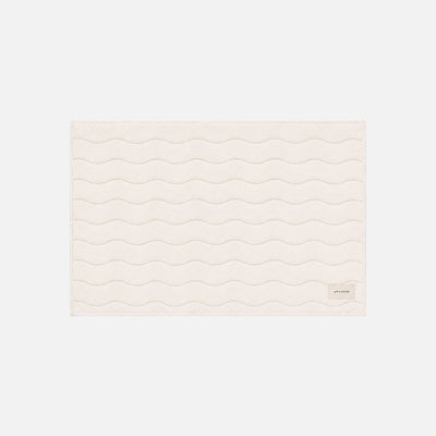 Cotton bath mat, cream, with wavy stitching and small corner tag.