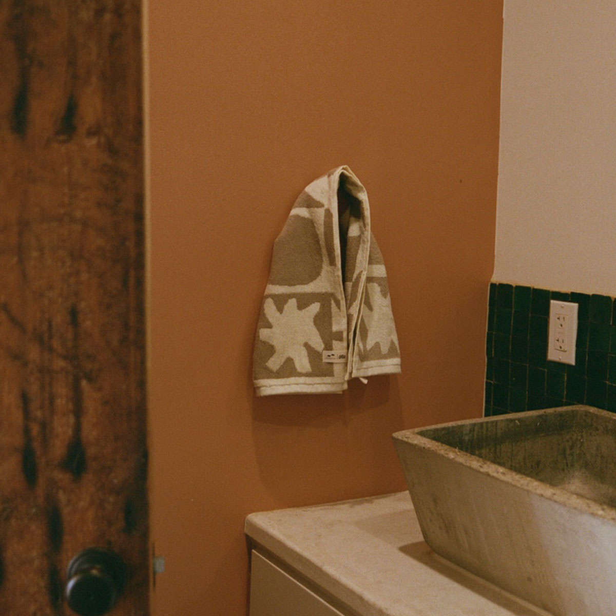 Bath towel with white flower pattern hanging on wall near green tiles.