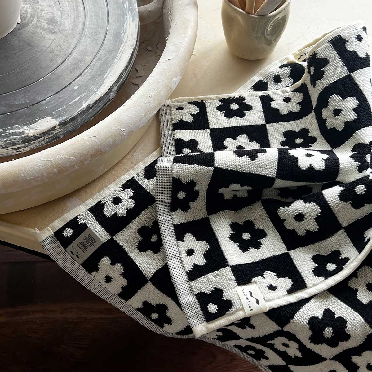 Cotton towel with black and white floral pattern next to pottery wheel and ceramic cup.