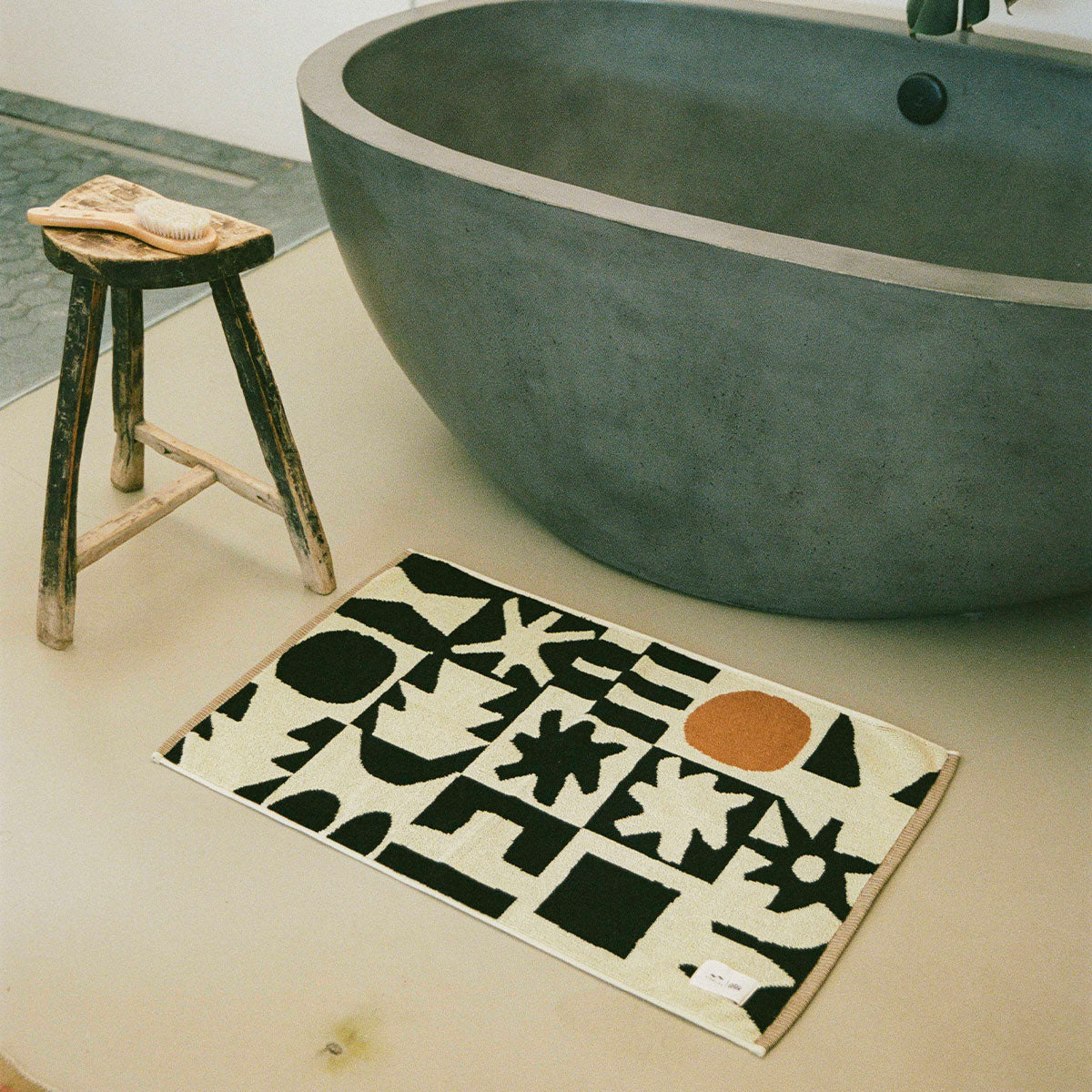 Concrete bathtub with absorbent bath mat and wooden stool in bathroom setting.