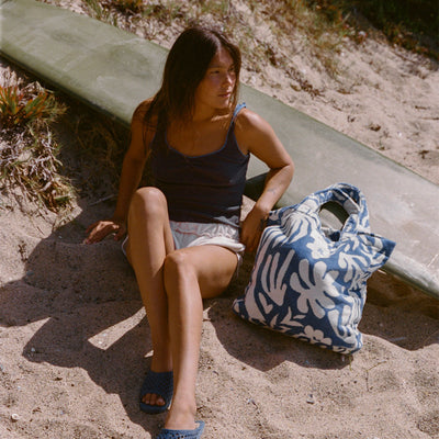 Large blue-and-white floral tote bag with surfboard on sandy beach.